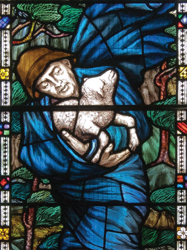 Stained glass depicting a figure in blue holding a lamb, surrounded by trees and colorful geometric patterns