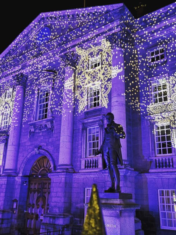 Trinity College illuminated with vibrant purple lights and intricate patterns, featuring a statue in the foreground