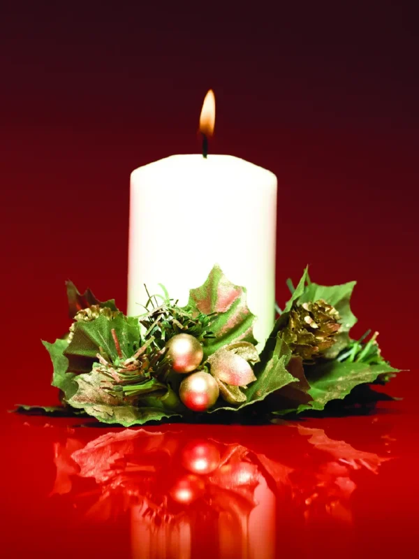 A lit white candle surrounded by green foliage and ornaments, reflecting on a red surface, evokes a festive mood for The Whit